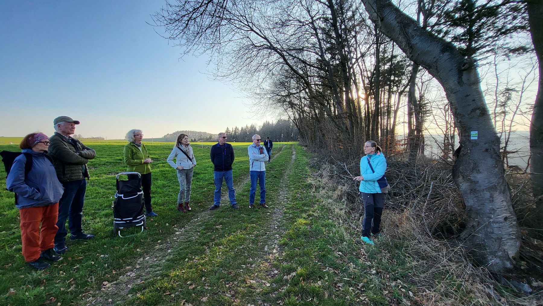 A group of seven people stand on a grassy trail at the edge of a field, listening to a woman who is speaking near a tree. Trees line one side of the path, and the sky is clear with sunlight filtering through.